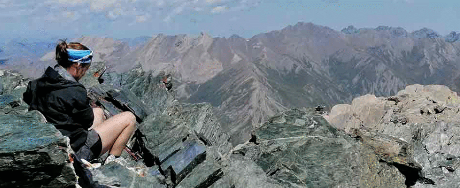 Una donna con una camicia nera e pantaloni corti è seduta di lato su rocce affilate su una montagna. La persona indossa un fascia sulla testa e guarda verso un paesaggio montuoso. Le vette delle montagne si estendono in lontananza sotto un cielo azzurro. Il terreno è aspro e roccioso, e la scena trasmette un senso di altezza e tranquillità.
