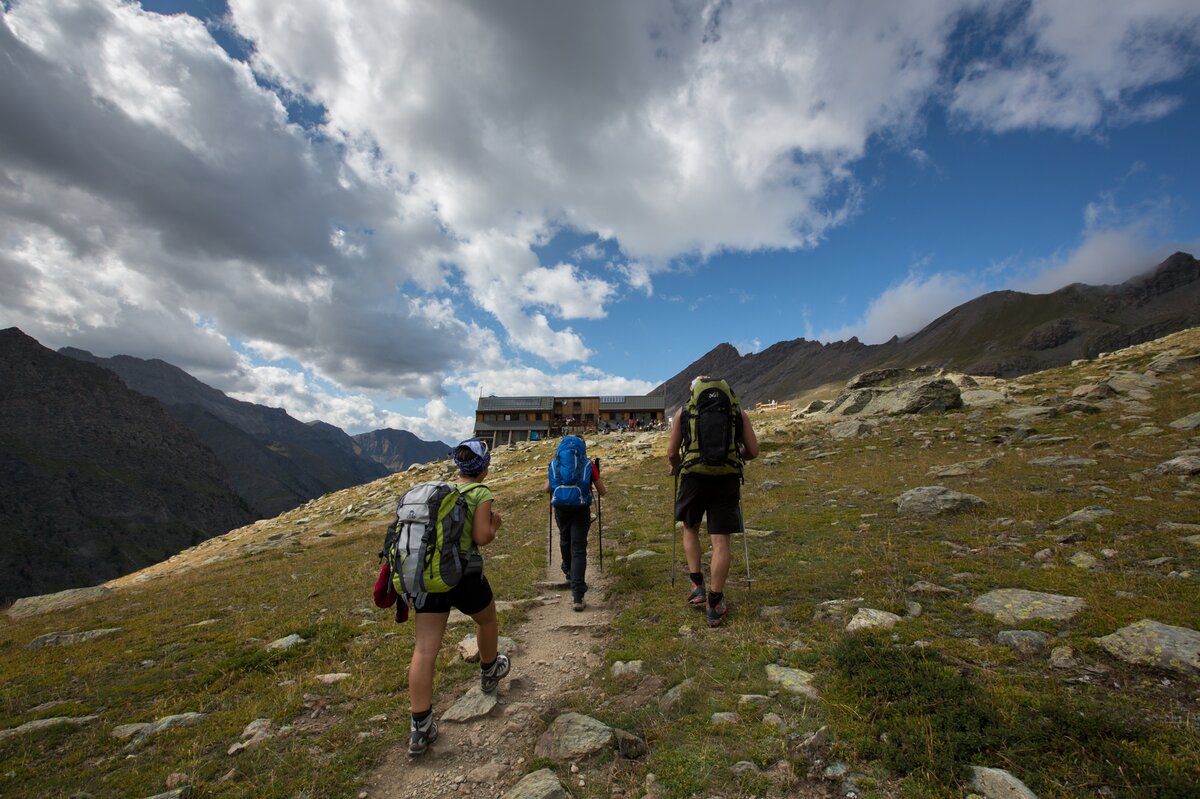 tre escursionisti su un sentiero, di spalle. di fronte a loro un rifugio a breve distanza.