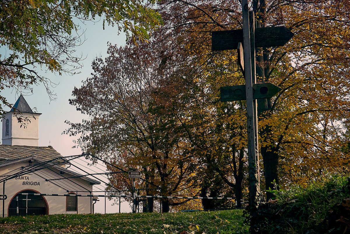 la facciata della cappella di Santa Brigida colta dallo spiazzo antistante, circondato da alberi e con due paline informative degli itinerari escursionistici che passano in loco.