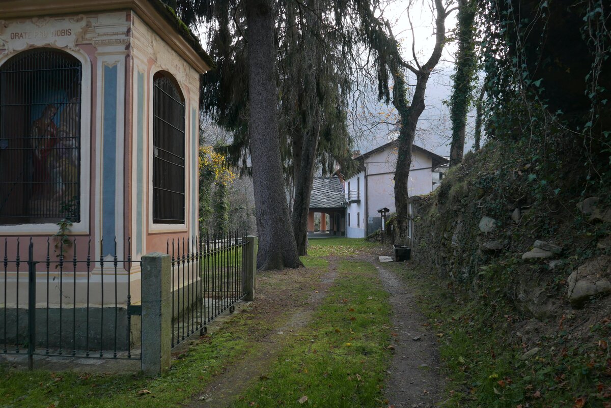 in primo piano sulla sinistra un pilone votivo, sullo sfondo al centro della foto e al fondo di un sentiero alberato il Santuario, visto lateralmente