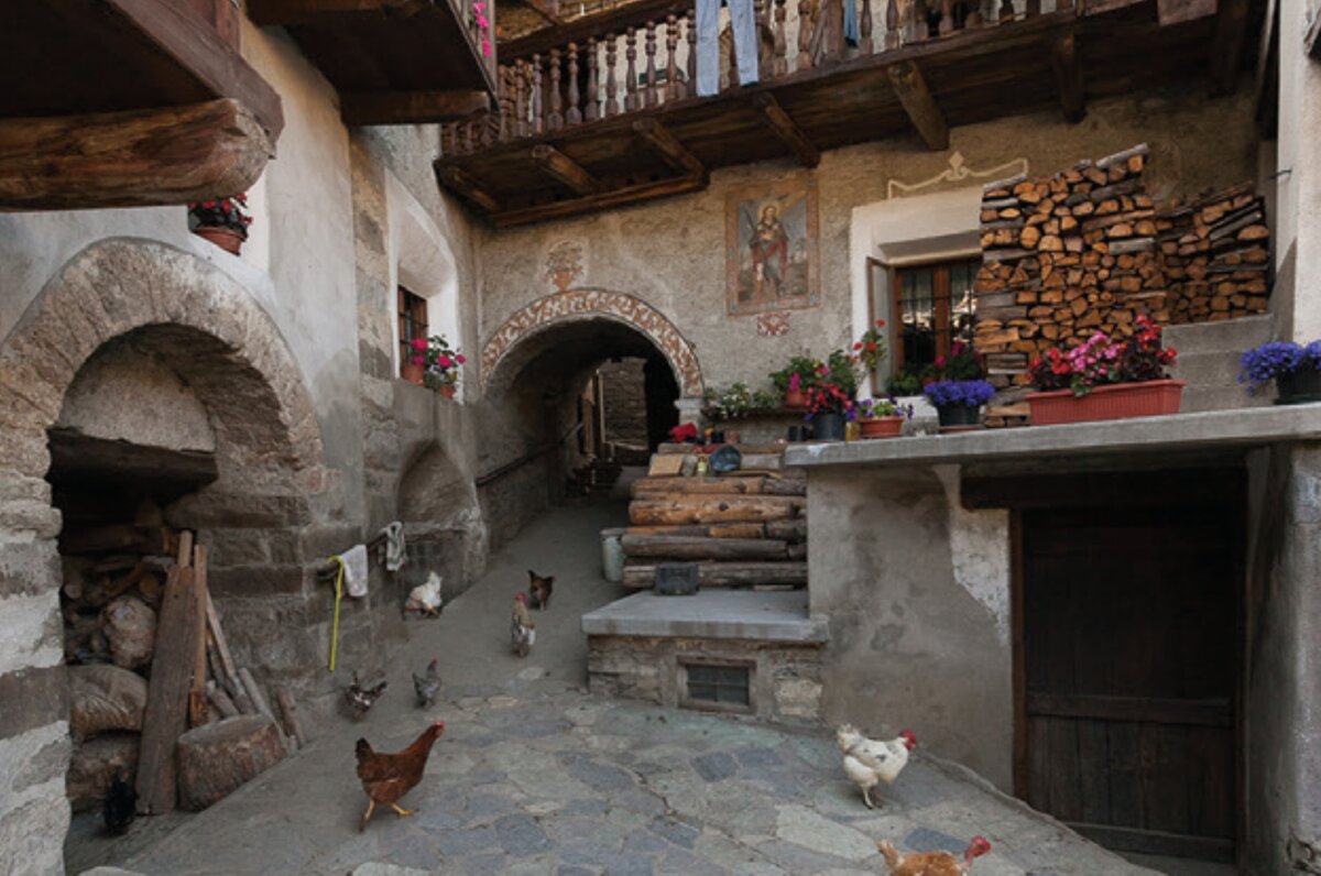 uno scenario di borgata, con una corte nella quale ci sono alcune galline, legname, balconi e alcune decorazioni pittoriche