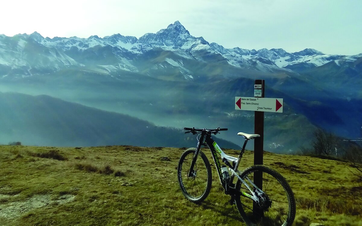 una bicicletta MTB appoggiata ad una palina escursionistica; sullo sfondo il Monviso e la sua catena.