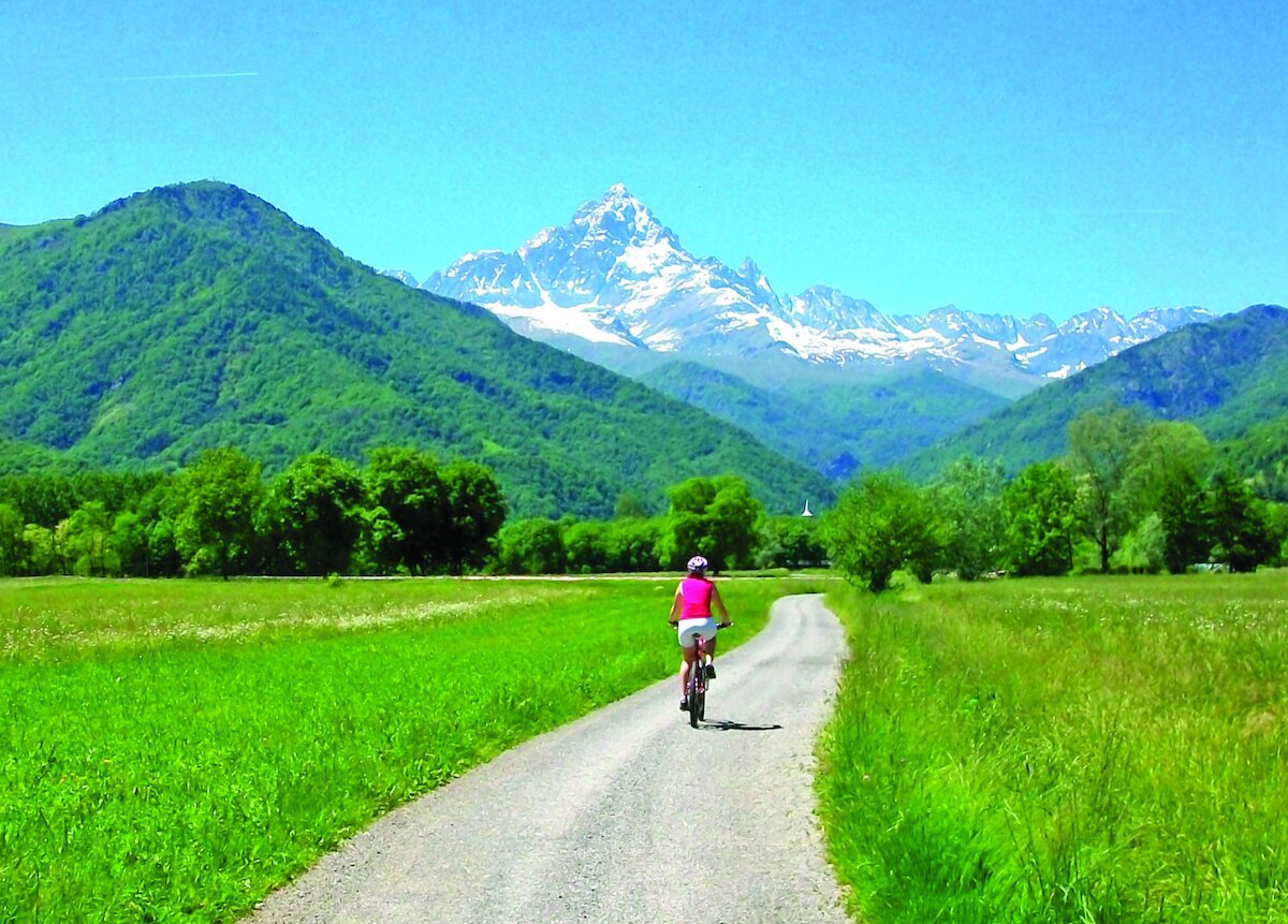 una ciclista di spalle sta percorrendo una strada asfaltata a viabilità secondaria, sullo sfondo e nella direzione verso la quale si sta dirigendo si staglia il Monviso