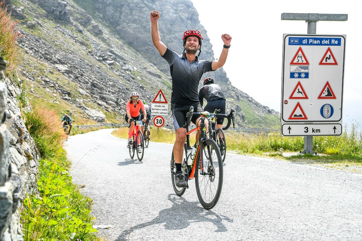 un ciclista sta risalendo la strada asfaltata per il Pian del Re e, essendo vicino all'arrivo, alza le braccia al cielo per esultare.