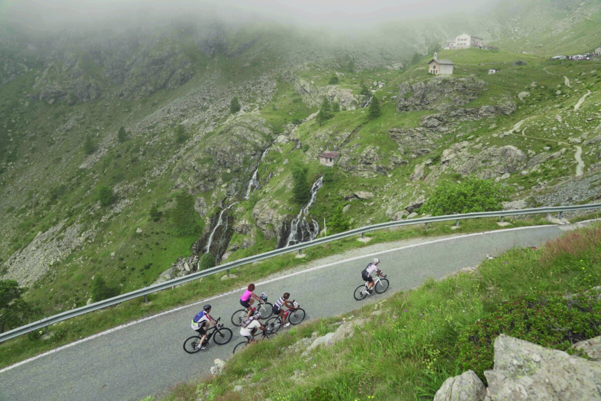 alcuni ciclisti risalgono la strada per il Pian del Re in una giornata con leggera foschia. In alto nella foto si vede la cappella del Pian del Re e la cascatella sottostante.