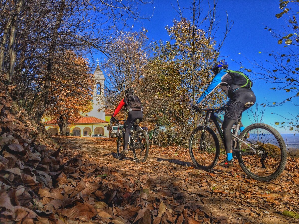 due ciclisti si avvicinano alla cappella: il fondo del sentiero è coperto da foglie cadute, il cielo è sereno.
