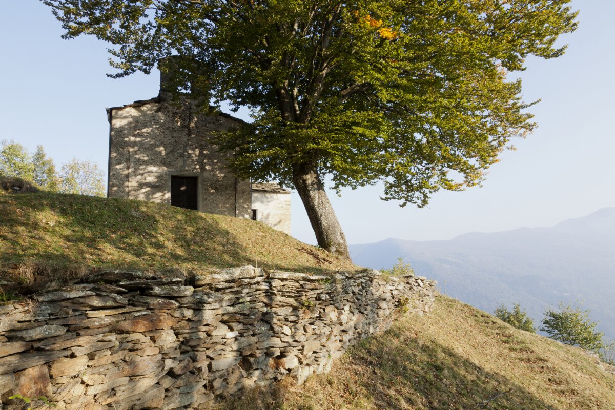 una cappella posta in un luogo panoramico sulla bassa valle: l'edificio si trova a sinistra nell'immagine e davanti ad esso c'è un albero frondoso